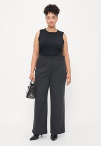Schwarzes ärmelloses Oberteil, graue maßgeschneiderte hochtaillierte Hose mit seitlichen Knopfdetails, schwarze spitze Schuhe und eine kleine schwarze Handtasche.