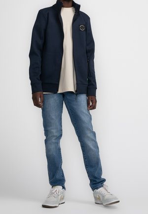 Modrá zipová bunda s vysokým límcem, béžové tričko a světle modré džíny. Tenisky bílé barvy s béžovými akcenty. Neformální design oblečení.