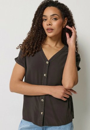 Kvinde med krøllet hår iført en sort kortærmet skjorte med knapper og lyseblå jeans, som poser med den ene hånd nær ansigtet.