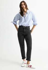 Giovane donna che indossa una camicia azzurra a righe con bottoni, jeans neri a vita alta con cintura e sneakers bianche con dettagli rossi, in piedi.