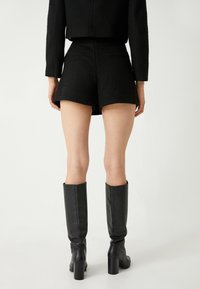 Completo corto nero strutturato con vita aderente, abbinato a stivali neri con tacco al ginocchio. L'outfit è presentato di spalle.