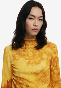 Gelbes Langarmoberteil mit floralem Muster und strukturiertem Stoff. Hochgeschlossener Schnitt mit taillierter Silhouette. Glatte Oberflächendetails.