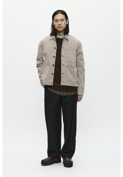 Beige canvas jasje met voorzakken, bruine gebreide trui en geruit shirt, gecombineerd met zwarte broek en bruine veterschoenen.