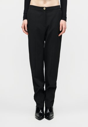 Pantalon noir ajusté à jambe droite avec pli sur le devant, fermeture par bouton doré, porté avec des bottines noires à talons pointues et un top noir court.