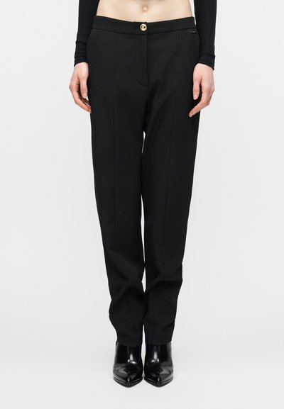 Pantalon noir ajusté à jambe droite avec pli sur le devant, fermeture par bouton doré, porté avec des bottines noires à talons pointues et un top noir court.