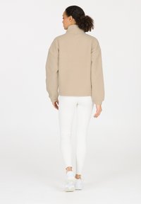 Beige Fleecejacke mit hohem Kragen, oversized Passform und gerippten Bündchen. Kombiniert mit weißen Leggings und hellen Sneakern.