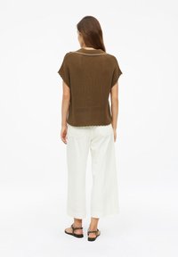 Mujer con cabello largo y castaño que lleva un top de punto marrón de manga corta, pantalones blancos de pierna ancha y sandalias negras, de pie de espaldas.