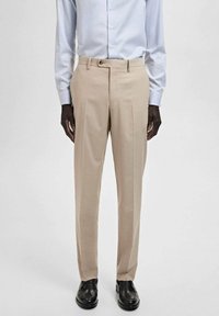 Beige dress pants with sharp creases, tailored fit, belt loops, and a smooth texture. Paired with a light blue button-up shirt and black shoes.