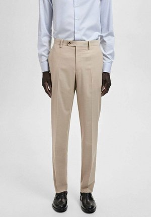 Pantaloni beige con pieghe nette, vestibilità tailored, passanti per la cintura e una texture liscia. Abbinati a una camicia button-up azzurro chiaro e scarpe nere.