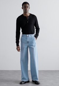 Camisa henley negra de manga larga combinada con pantalones vaqueros de pierna ancha azul claro. Los vaqueros tienen una cintura alta y un cierre con botón, complementados por zapatos negros brillantes.