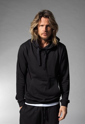 Schwarzer Kapuzenpullover mit langen Ärmeln, Kängurutasche und Kordelzug. Weicher Stoff, lässige Passform, kombiniert mit schwarzen Hosen.
