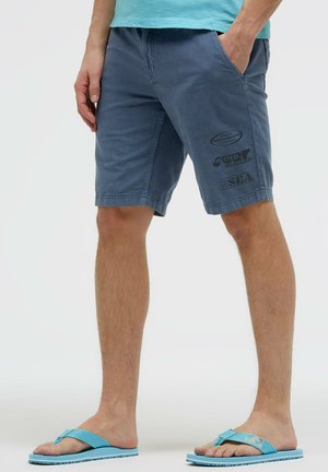 Man wearing blue shorts with printed text on left thigh, teal shirt, and blue flip-flops, standing with hands in pockets.