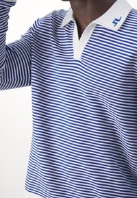 Raido pitkähihaine polo paita sinisen ja valkoisen raidallisen puuvillan sekoituksesta, jossa on valkoinen kaulus ja sininen logon yksityiskohta kauluksessa.