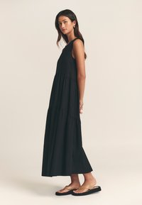 Juoda maxi suknelė su sluoksniuota konstrukcija, be rankovių ir minkšta faktūra. Derinama su plokščiais, auksiniais akcentais puoštomis šlepetėmis.