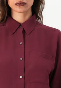 Femme portant une chemise bordeaux boutonnée avec un col pointu et une poche poitrine, visible du menton à l'épaule sur fond blanc.
