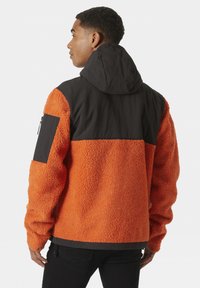 Fleece bunda s černou kapucí a horním panelem, s oranžovým texturovaným tělem, dlouhými rukávy a bočním zipovým kapsou.