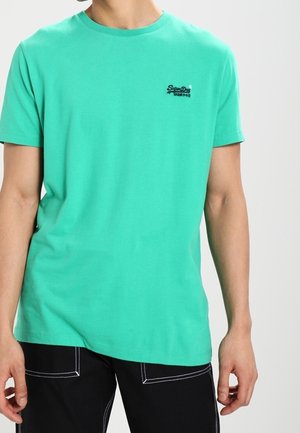 Man draagt een effen turquoise shirt met korte mouwen, met een klein zwart logo op de linkerborst, en zwarte broek met witte stiksels.