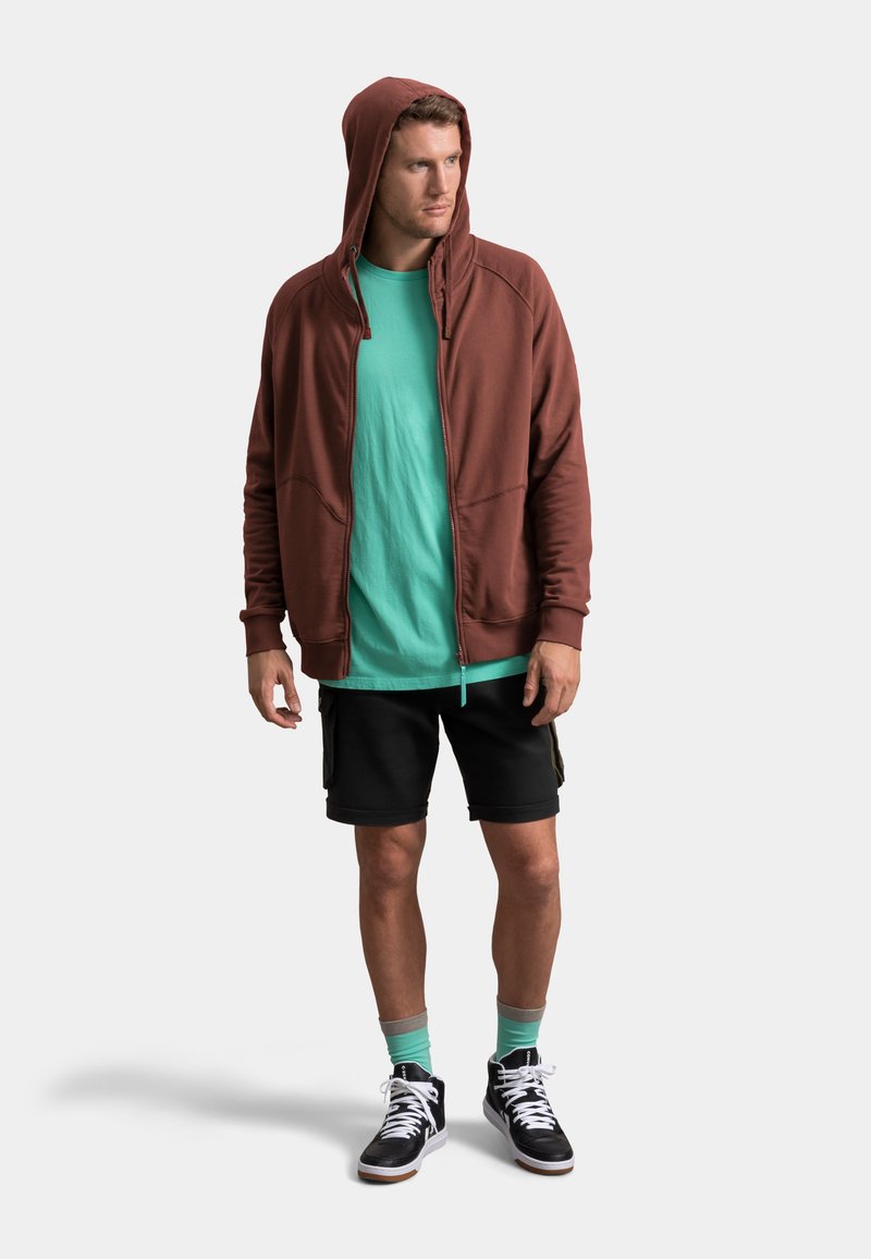 Felpa marrone con zip, maglietta verde, pantaloni cargo neri e sneakers alte. Completato da calzini verdi e un design minimalista.