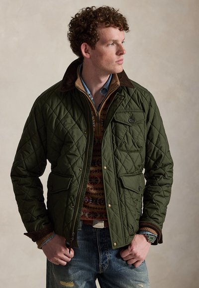 Młody mężczyzna z kręconymi włosami, ubrany w zieloną pikowaną kurtkę, sweter we wzory, jeansową koszulę i niebieskie jeansy, patrzący w prawo.