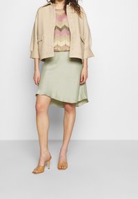 Light beige cropped jacket, patterned knitted top with chevron design in pink and green, flowing green satin skirt, and tan heeled sandals.