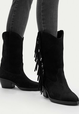 Schwarze Wildleder-Stiefel bis zur Mitte der Wade mit Fransen, kombiniert mit grauen, eng anliegenden Jeans, niedrigen gestapelten Absätzen und spitzen Zehen.