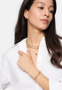Set di gioielli in oro composto da catene sovrapposte, un piccolo pendente e orecchini a bottone rotondi. Il modello indossa una camicia bianca con bottoni.