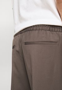 Detailní záběr na šedé elastické kalhoty s kapsami na zadní straně, které jsou nošeny s bílou košilí zasunutou uvnitř.