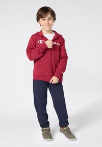 Junge in rotem Champion-Hoodie, blauen Hosen und olivgrünen Champion-Sneakers, stehend und lächelnd mit der Hand nahe dem Reißverschluss.