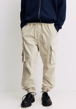 Beige Cargohosen mit elastischen Bündchen, mehreren Seitentaschen und einem Tunnelzugbund, kombiniert mit schwarzen Schnürschuhen.