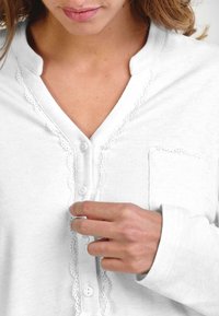 Mujer abrochando los botones de una blusa blanca de manga larga con encaje y bolsillo en el pecho, mostrada desde el cuello hasta la mano.