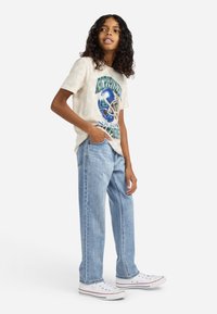 Niño con pelo rizado que lleva una camiseta gráfica beige, jeans azul claro y zapatillas blancas, de pie con una mano en el bolsillo.