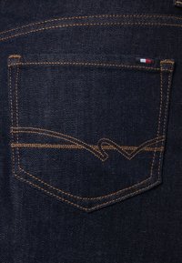 Bolsillo de mezclilla oscura con costura amarilla contrastante y un diseño ondulado. Pequeña etiqueta de marca con colores rojo, blanco y azul en el borde del bolsillo.