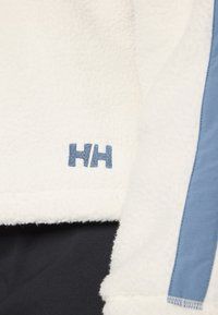 Közelkép egy fehér bolyhos ruhadarabról, amelyen kék függőleges csík található, és az "HH" logo kék szálakkal belevarrva a szövetbe.