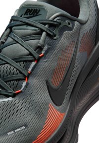 Zapatilla de running Nike gris con parte superior texturizada, símbolo negro, detalles naranjas y una lengüeta acolchada etiquetada como "RUN". Cuenta con cordones negros y suela duradera.