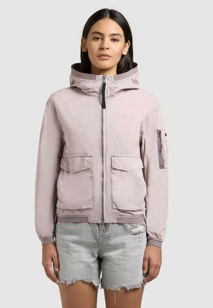 Młoda kobieta w jasnoróżowej kurtce z kapturem i kieszeniami z patkami oraz jasnoszarych, postarzanych krótkich spodniach z denimu, stojąca na tle jednolitego tła.