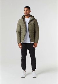 Casaco tipo puffer verde-azeitona com capuz; textura canelada; logotipo preto na manga. Combinado com calças cargo pretas e ténis brancos.