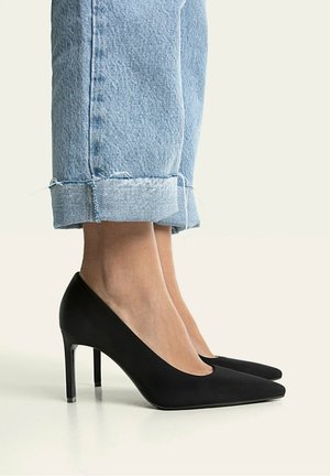 Feet wearing black pointed-toe high heels paired with light blue cuffed jeans against a plain light background.