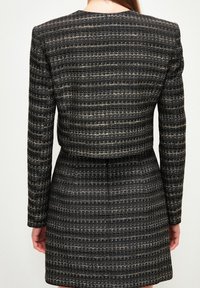 Vrouw in een nauwsluitende, langmouwige, zwarte jurk met horizontale witte en metallic strepen, gezien vanaf de achterkant tegen een effen achtergrond.