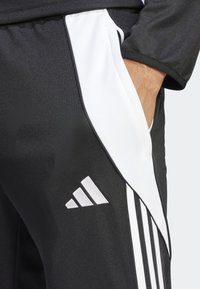 Svarta träningsbyxor med vita sidosträck och en Adidas-logotyp. Tillverkade av ett slätt tyg med en texturerad yta och en ficka med dragkedja på sidan.