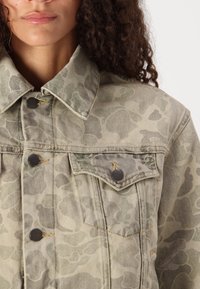 Kamouflage-mønstret denimjakke i dæmpede grønne og beige nuancer. Har en skjortekrave, frontknapper og brystlommer med sort metaludstyr.