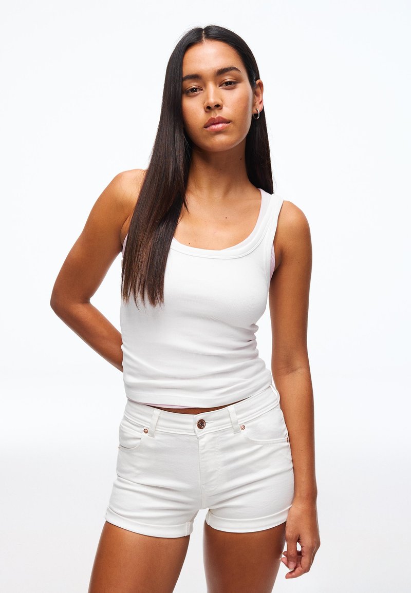 Giovane donna con capelli lunghi e scuri, indossa una canottiera bianca e pantaloncini di jeans bianchi, in piedi con una mano sul fianco contro uno sfondo bianco.