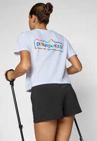 T-shirt gris clair à manches courtes avec un logo coloré "Patagonia" au dos ; associé à un short noir et tenant une canne de marche.