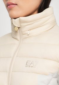Beige pufferjack met een hoge kraag en horizontale stiksels. Voorzien van gouden hardware en een glanzend logo op de borst. Voorzien van een gladde textuur.