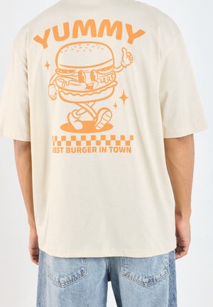 Beżowy t-shirt z pomarańczową grafiką kreskówkowego burgera pokazującego kciuk w górę, napis „YUMMY” u góry oraz „BEST BURGER IN TOWN” na dole.
