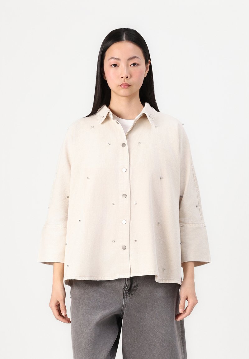Camicia beige con bottoni, dal taglio rilassato, decorata con piccole decorazioni in argento. Presenta un colletto e maniche ampie.