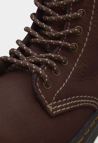 Κοντινή λήψη καφέ δερμάτινου μποτάκι με λευκές ραφές, μεταλλικά χάντρες, και χοντρά καφέ κορδόνια με λευκές λεπτομέρειες.