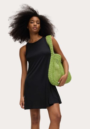 Kobieta z kręconymi włosami, ubrana w czarną, bezrękawną sukienkę, trzymająca dużą zieloną, dzianą torbę na ramię na jednolitym tle.
