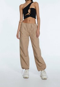 Top corto negro de un solo hombro con detalle de corte, combinado con pantalones beiges holgados con cintura elástica y cordón, usados con zapatillas blancas.