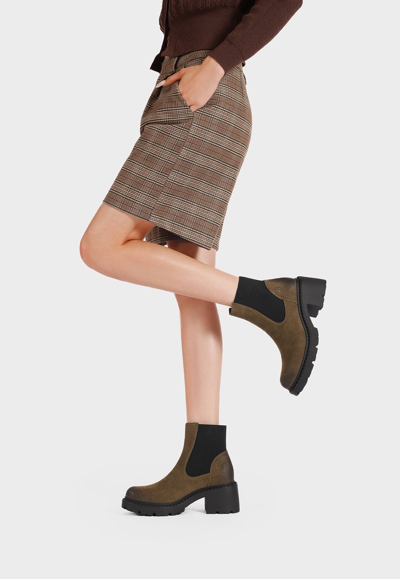 Stivaletti alla caviglia in pelle scamosciata marrone con pannelli elastici laterali, punte quadrate e suole in gomma nere spesse, abbinati a una gonna a quadri.