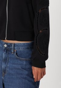 Čierny skrátený sweatshirt s predným zipsom, textúrovanými detailmi na rukávoch a rebrovanými manžetami, doplnený o vysoké modré denimové džínsy.